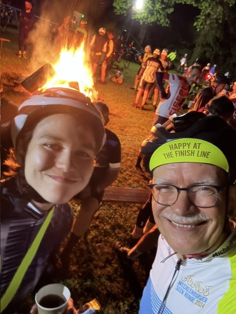 Ein kleines Lagerfeuer brennt unter einem großen Baum, im Hintergrund weitere Zelte und Fahrzeuge.